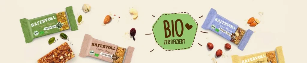 Vier Hafervoll Organic Flapjack Riegel verschiedener Sorten mit Bio-Zertifiziert-Siegel und Zutaten auf hellem Hintergrund