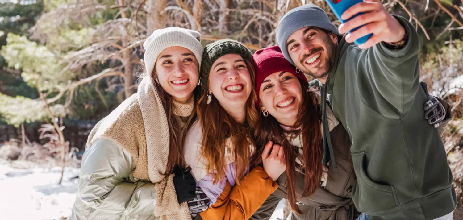 Vier lachende junge Menschen machen ein Selfie im verschneiten Wald mit Wintermützen