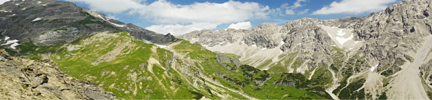 Panoramablick auf grüne Almwiesen und felsige Alpengipfel mit Schneeresten unter blauem Himmel